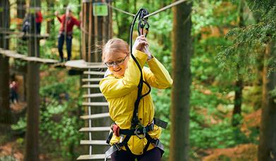 Menyw mewn cot law felen yn dringo ar draws rhwystr yn Go Ape Margam