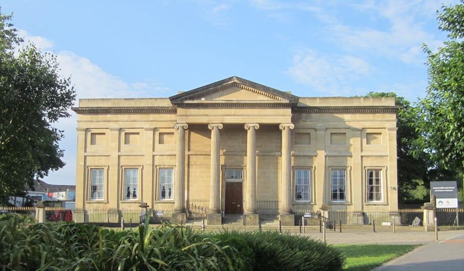 View of front of museum a classical building in light sand coloured stone. The photograph shows six large windows and four ionic colums by the front e