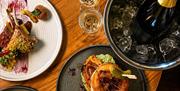 Overhead view of dishes on a table. Gower butcher beef burger, Rack of Gower lamb and Gower Gold beer battered cod and chips. Bottle of Prosecco.