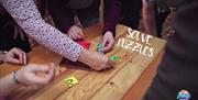 A large wooden box with colourful jigsaw pieces and several pairs of hands trying to solve the clue. SOLVE PUZZLES overlaid